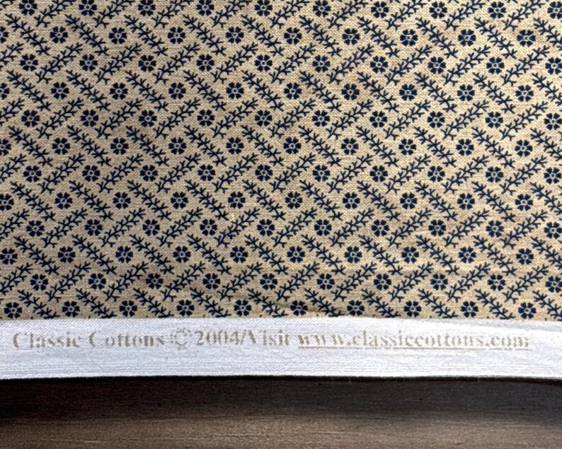 Puede incluir: Primer plano de una tela con un patr&oacute;n repetido de motivos florales y de vid azul marino sobre un fondo beige. La tela tiene un dise&ntilde;o diagonal. El texto "Classic Cottons" y una direcci&oacute;n web son visibles en la parte inferior.