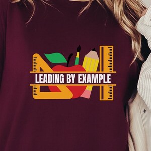 May include: A maroon t-shirt with a graphic of a red apple, pencil, ruler, and protractor. The text "LEADING BY EXAMPLE" is printed on the shirt.