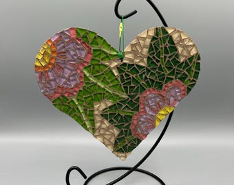 May include: A heart-shaped mosaic art piece with a floral design in shades of green, purple, yellow, and beige. The mosaic is displayed on a black metal stand with a circular base and a curved top.