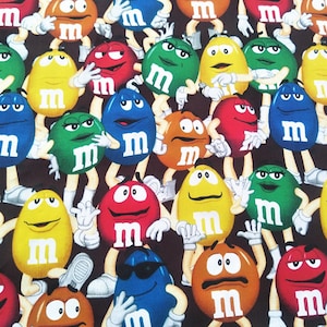 Op de afbeelding: Stof met een herhalend patroon van kleurrijke M&M's snoepjes. De snoepjes zijn in verschillende kleuren, waaronder rood, geel, groen, blauw en oranje, elk met een witte "m" op de voorkant. De achtergrond is donkerbruin.