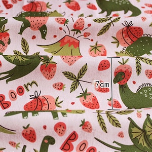May include: A white fabric with a repeating pattern of green dinosaurs and red strawberries. The strawberries have green leaves and are in various sizes. The word "Boom" is visible in the lower left corner. The measurement is 7cm.