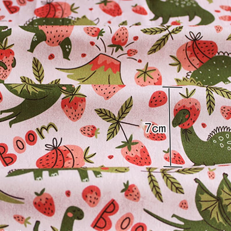 May include: A white fabric with a repeating pattern of green dinosaurs and red strawberries. The strawberries have green leaves and are in various sizes. The word "Boom" is visible in the lower left corner. The measurement is 7cm.