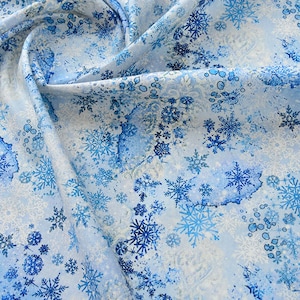 May include: A light blue fabric featuring a repeating pattern of dark blue and white snowflakes in various sizes. The design includes watercolor-style blue splatters and dots scattered across the surface, creating a layered winter-themed textile.
