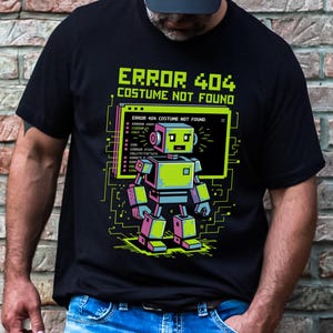 Könnte beinhalten: Schwarzes T-Shirt mit einem neongrünen und pinkfarbenen Roboter-Grafik und dem Text "ERROR 404 COSTUME NOT FOUND". Das Roboter-Design hat einen pixeligen Stil mit Leiterplattenelementen.