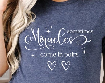 May include: A navy blue t-shirt with the text "Miracles sometimes come in pairs" in white lettering. Two white hearts are below the text.