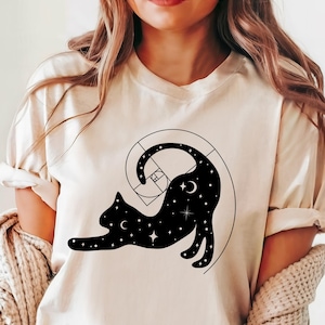 May include: A white t-shirt with a black silhouette of a cat curled up in a spiral. The cat is filled with stars and crescent moons.