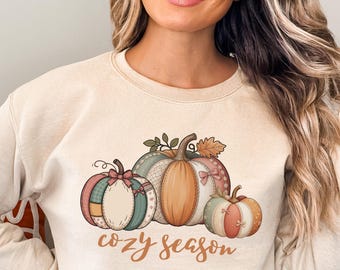 Op de afbeelding: Crèmekleurige sweatshirt met een herfstthema van drie pompoenen in verschillende kleuren en patronen. De zin "cozy season" is in cursieve letters onder de pompoenen gedrukt. De sweatshirt heeft een ronde hals en lange mouwen.