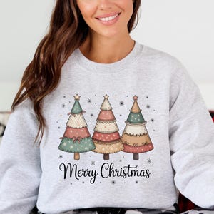 May include: A light grey sweatshirt with three pastel-coloured Christmas trees. The trees are decorated with stars and ornaments, with the text "Merry Christmas" underneath. The sweatshirt is a comfortable, casual style.