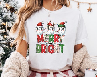 May include: White t-shirt with a Christmas-themed graphic. The design features three cartoon teeth wearing Santa hats and string lights, with the words "Merry and Bright" in festive lettering. The person is wearing red and white plaid pajama pants.
