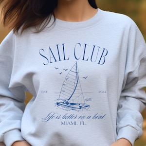 May include: A light blue sweatshirt with a graphic design of a sailboat and the text "SAIL CLUB EST. 2024 Life is better on a boat MIAMI, FL"