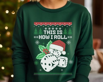 May include: A dark green sweatshirt with a festive design. The design includes the text "THIS IS HOW I ROLL" with dice wearing a Santa hat. The design also features snowflakes, Christmas trees, and a red and white patterned border.