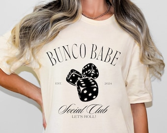 May include: A cream-colored t-shirt with black text that reads "Bunco Babe Est. 2024 Social Club Let's Roll!" and a graphic of three black dice.