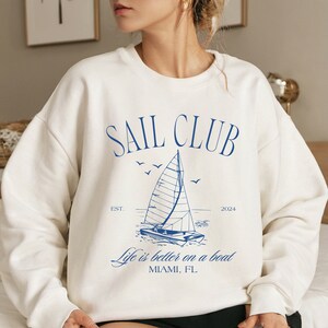 May include: White sweatshirt with a blue graphic of a sailboat and the text "SAIL CLUB EST. 2024 Life is better on a boat MIAMI, FL"