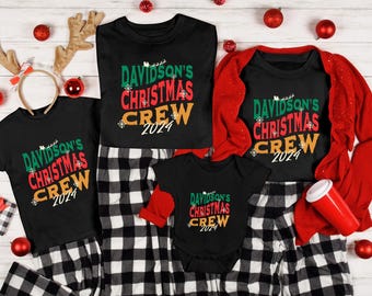 May include: A family Christmas pajama set featuring black shirts and a onesie with the text "Davidson's Christmas Crew 2024" in green, red, and orange. The set includes matching black and white checkered pajama pants and a red long-sleeved top.