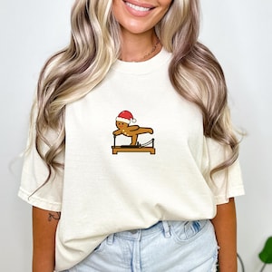 May include: Cream-colored t-shirt featuring a gingerbread man wearing a Santa hat, exercising on a Pilates reformer. The gingerbread man is brown with black details. The reformer is brown with black and silver accents. A fun, festive design.