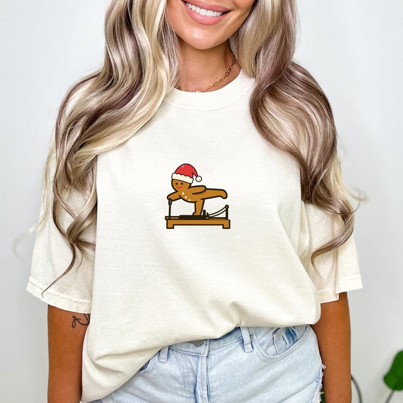May include: Cream-colored t-shirt featuring a gingerbread man wearing a Santa hat, exercising on a Pilates reformer. The gingerbread man is brown with black details. The reformer is brown with black and silver accents. A fun, festive design.
