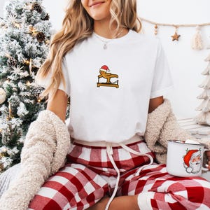 May include: White t-shirt featuring a gingerbread man wearing a Santa hat, doing gymnastics on a brown apparatus. The image also shows red and white plaid pajama pants and a mug with a Santa Claus design.