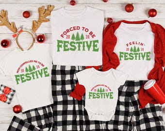 May include: White t-shirts and a baby onesie with green text that reads "FESTIVE" and "20 24" with Christmas tree graphics. The shirts are paired with black and white plaid pajama pants. Red Christmas ornaments and reindeer antlers are scattered around the shirts.