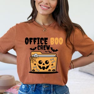 May include: A burnt orange t-shirt with the words "OFFICE BOO CREW" in black and orange. The graphic features a Halloween-themed file folder with a jack-o'-lantern face, spiderwebs, bats, and bones. A fun and festive design for Halloween.