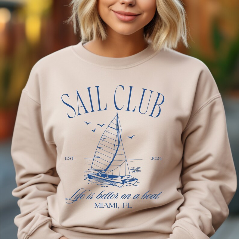 May include: A beige sweatshirt with a blue graphic design of a sailboat and the text "Sail Club Est. 2024 Life is better on a boat Miami, FL"