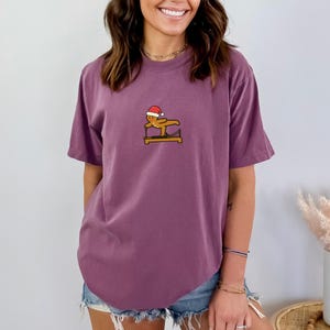 May include: A mauve-colored t-shirt featuring a small graphic of a gingerbread man wearing a Santa hat, balancing on a balance beam. The gingerbread man is brown with a red and white hat. The shirt has short sleeves and a relaxed fit.