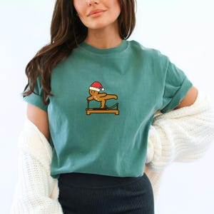 May include: Teal-colored t-shirt featuring a gingerbread man wearing a Santa hat, performing a Pilates exercise on a wooden apparatus. The graphic is in the center of the shirt. The shirt has short sleeves and a relaxed fit.