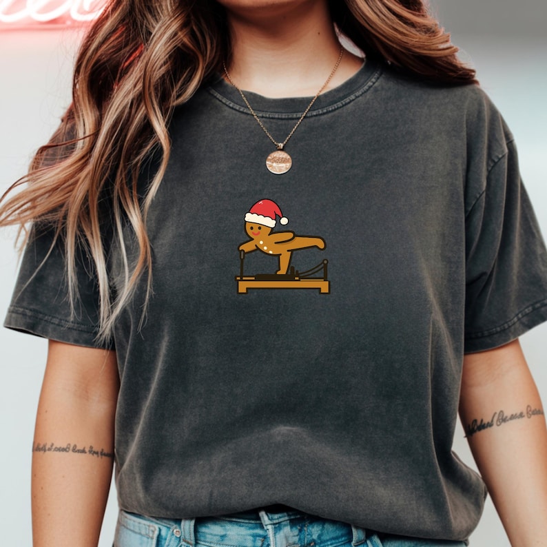 May include: Dark gray t-shirt featuring a gingerbread man wearing a Santa hat, exercising on a Pilates reformer. The graphic is in shades of brown, red, and white. The shirt is a casual style, suitable for everyday wear.