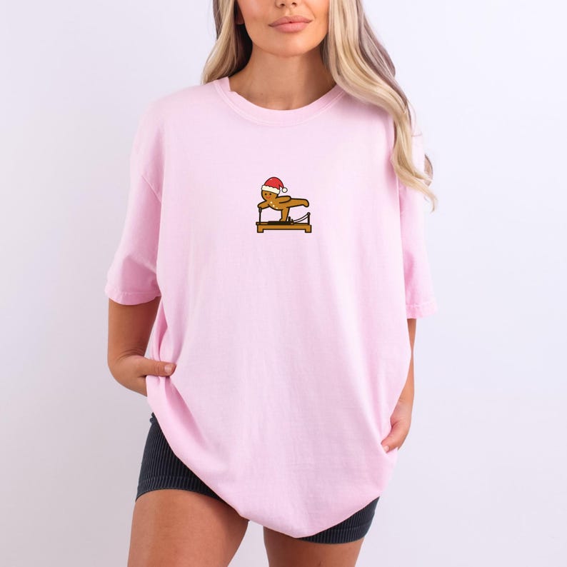 May include: A light pink t-shirt featuring a small graphic of a gingerbread man wearing a Santa hat, balancing on a wooden beam. The shirt has short sleeves and a relaxed fit.