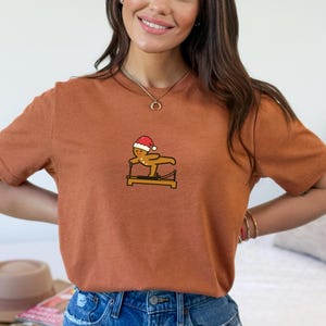 May include: A rust-colored t-shirt featuring a cartoon gingerbread man wearing a Santa hat and exercising on a Pilates reformer. The design is centered on the chest. The shirt has short sleeves.