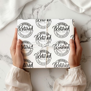 May include: A gift wrapped in white paper with a repeating black circular design that reads "Officially Retired est. 2025. It's not your problem anymore." The gift is tied with white ribbon and held by a person.