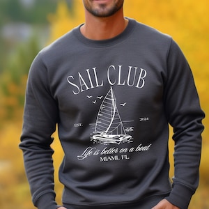 May include: A dark grey sweatshirt with the text "SAIL CLUB" in white, a sailboat graphic, and the text "Life is better on a boat" with the location "MIAMI, FL".