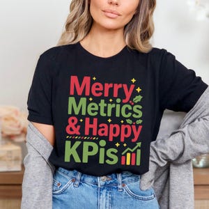 Pode incluir: Camiseta preta com as palavras "Merry Metrics & Happy KPIs" em vermelho e verde. O design inclui folhas de azevinho e detalhes de estrelas amarelas. A camiseta é feita de um material macio.