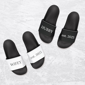 May include: Two pairs of black and white slides with the words "WIFEY" and "HUBBY" on one pair and "est. 2025" on the other pair.