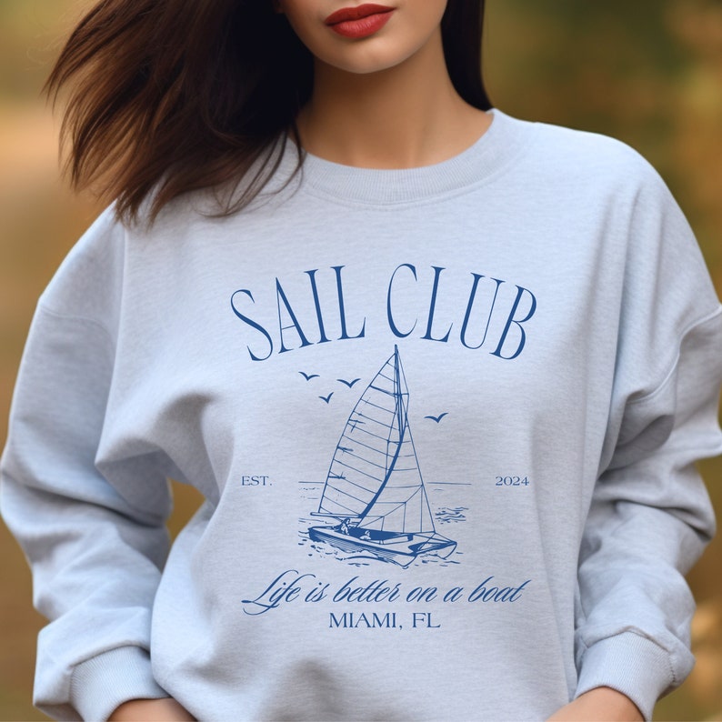 May include: A light blue sweatshirt with a graphic design of a sailboat and the text "SAIL CLUB EST. 2024 Life is better on a boat MIAMI, FL"