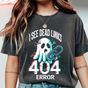 Può includere: T-shirt grigio scuro con un design grafico raffigurante un fantasma, un anello di catena rotto e il testo "I SEE DEAD LINKS 404 ERROR" con un effetto glitch. Il design utilizza colori bianco, blu e rosa.