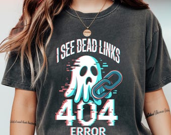 Może przedstawiać: Ciemnoszara koszulka z graficznym wzorem przedstawiającym ducha, przerwane ogniwo łańcucha oraz tekst "I SEE DEAD LINKS 404 ERROR" z efektem glitch. Projekt wykorzystuje kolory biały, niebieski i różowy.