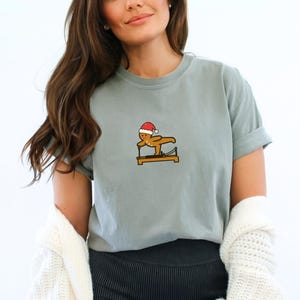 May include: A light blue t-shirt featuring a gingerbread man wearing a Santa hat, doing Pilates on a reformer machine. The graphic is in brown, red, and white. The shirt has short sleeves and a relaxed fit.