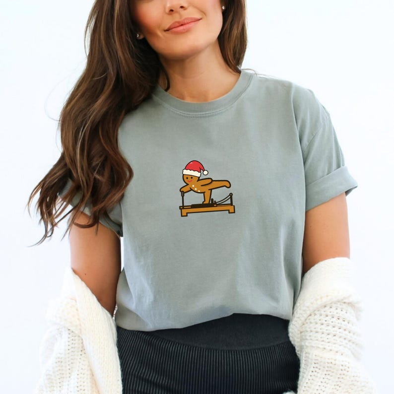 May include: A light blue t-shirt featuring a gingerbread man wearing a Santa hat, doing Pilates on a reformer machine. The graphic is in brown, red, and white. The shirt has short sleeves and a relaxed fit.
