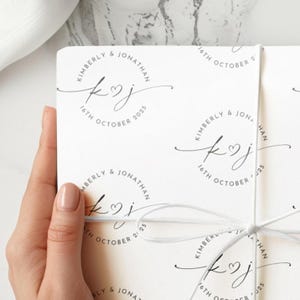 May include: White wedding stationery with a repeating circular design featuring the names "Kimberly & Jonathan", the date "16th October 2025", and initials "K♡J". The stationery is tied with a white ribbon.