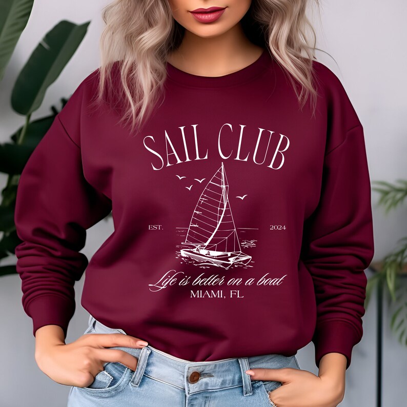 May include: A maroon sweatshirt with a white graphic design that says "Sail Club Est. 2024" with a sailboat and the text "Life is better on a boat Miami, FL"