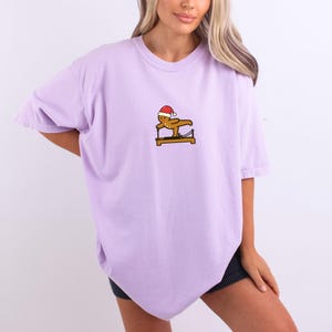 May include: A light purple t-shirt featuring a small graphic of a gingerbread man wearing a Santa hat, exercising on a Pilates reformer. The graphic is in shades of brown, red, and yellow. The shirt has short sleeves and a relaxed fit.
