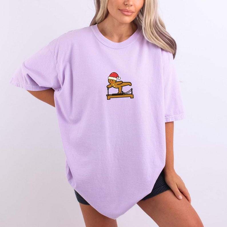 May include: A light purple t-shirt featuring a small graphic of a gingerbread man wearing a Santa hat, exercising on a Pilates reformer. The graphic is in shades of brown, red, and yellow. The shirt has short sleeves and a relaxed fit.
