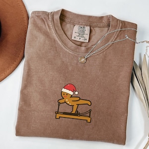May include: Brown Comfort Colors t-shirt with a crew neck and short sleeves. The shirt features a graphic of a gingerbread man wearing a Santa hat and exercising on a Pilates reformer. A silver necklace with a small pendant is draped across the shirt.