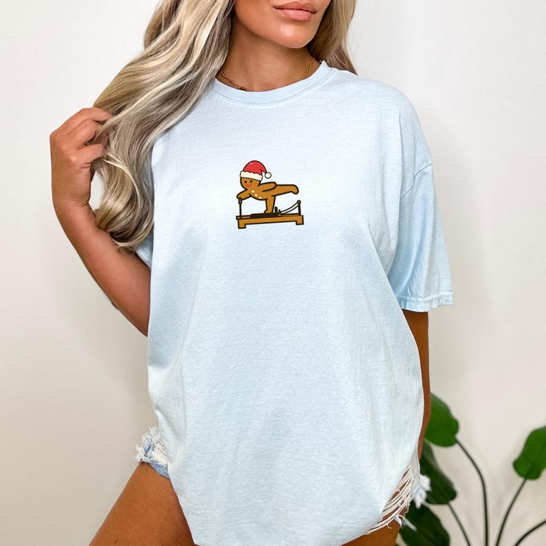 May include: Light blue t-shirt featuring a small graphic of a gingerbread man wearing a Santa hat, performing Pilates on a reformer. The shirt has short sleeves and a relaxed fit, suitable for casual wear.