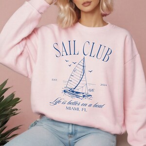 May include: Pink sweatshirt with a blue graphic of a sailboat and the text "SAIL CLUB EST. 2024 Life is better on a boat MIAMI, FL"