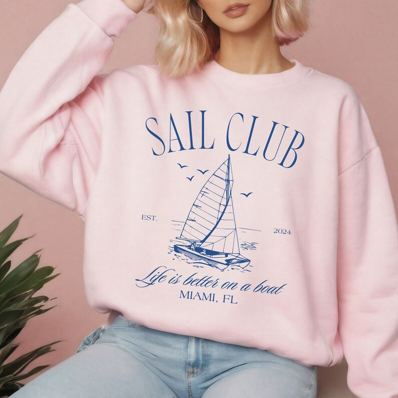 May include: Pink sweatshirt with a blue graphic of a sailboat and the text "SAIL CLUB EST. 2024 Life is better on a boat MIAMI, FL"