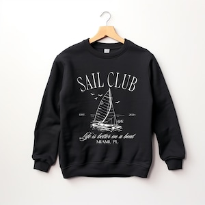 May include: Black sweatshirt with a white graphic design of a sailboat and the text "Sail Club Est. 2024 Life is better on a boat Miami, FL"