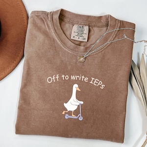 Könnte beinhalten: Braunes Comfort Colors T-Shirt mit dem Text "Off to write IEPs" und einer Cartoon-Gans auf einem Roller. Eine silberne Halskette mit Anhänger liegt über dem Shirt. Das Shirt hat die Größe S-P-CH.