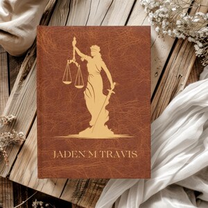 Puede incluir: Un diario de cuero marrón con una imagen dorada en relieve de la figura de la justicia sosteniendo una balanza y una espada. El diario tiene el nombre "Jaden M Travis" en relieve dorado en la portada.