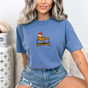 May include: A blue t-shirt featuring a gingerbread man wearing a Santa hat, exercising on a Pilates reformer. The graphic is in the center of the shirt. The shirt is made of a soft material.